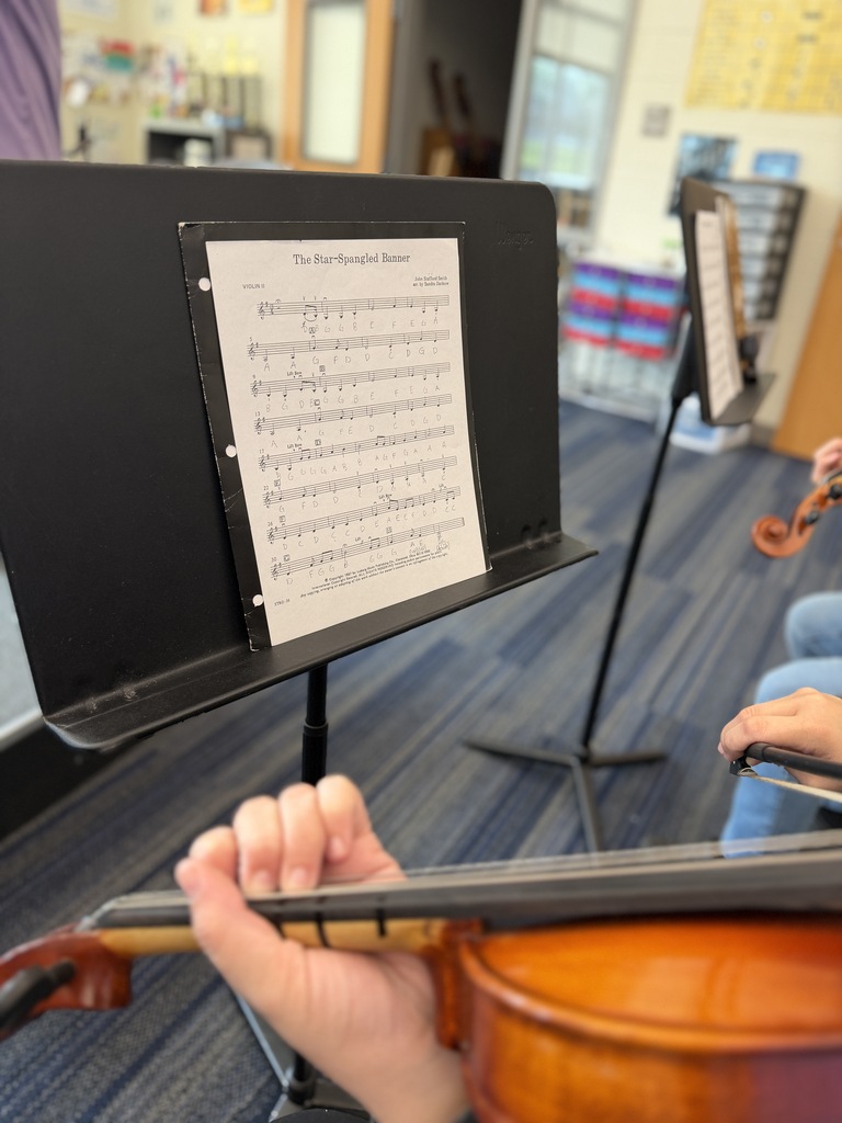 A music stand with the sheet music for The Star-Spangled Banner is shown resting on the stand. A student's hand is in the front part of the picture holding a violin.