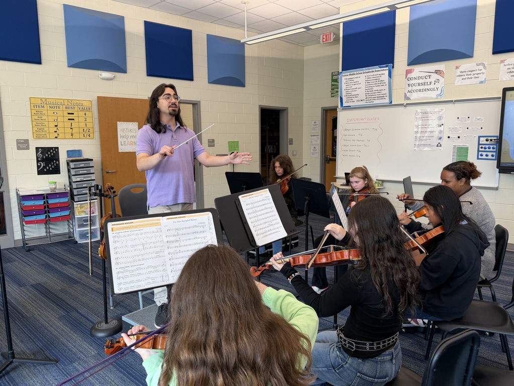 Students are seated in their chairs during 8th grade Orchestra classes with their instruments held in position and practicing The Star-Spangled Banner piece in preparation for the upcoming Veteran's Day Assembly. The Orchestra director is shown standing on the director's podium with a director stick in his hands guiding them through the song.