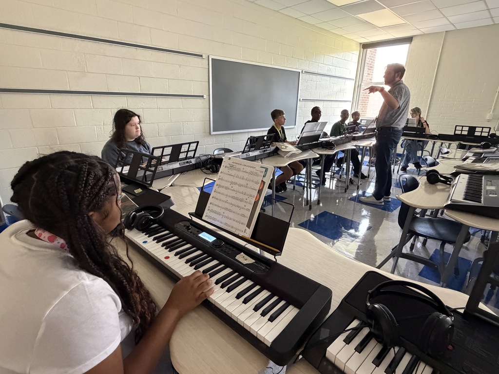 Percussion students are in a classroom with piano keyboards on desks.Their hands are resting on the keys while looking at their band instructor who is standing in the middle of a u-shaped row of desks sharing instructions.