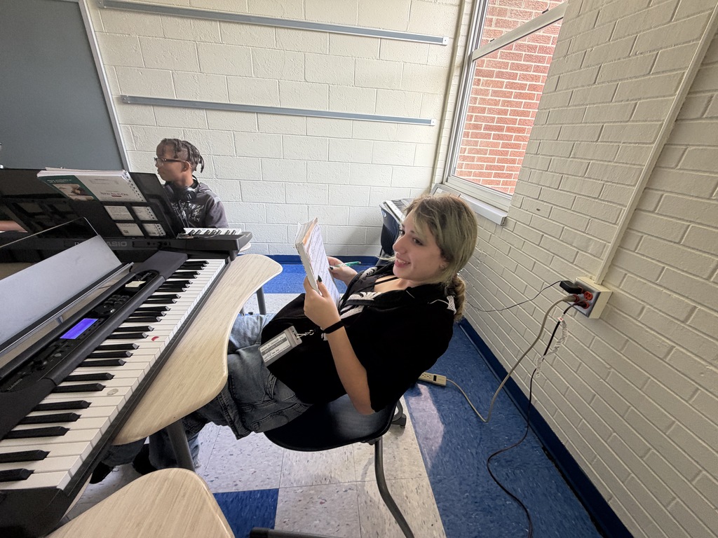 A female student is smiling while looking off at a different student in class. She is hold the sheet music and music book in her hands and leaning back in her chair. A male student is shown in the background seated at his desk.