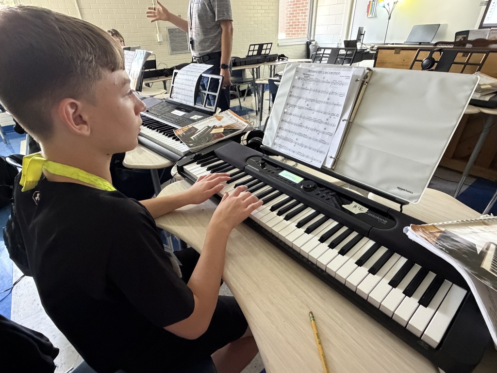 A male student is shown with his hands on the piano keys practicing certain keys while looking at his sheet music on the stand above the piano keyboard.