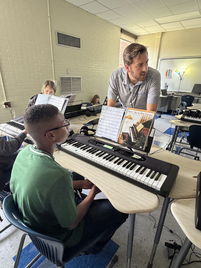 The band instructor is standing in front of a student's sheet music while the student is seated at his desk with the piano keyboard on his desk. The teacher is sharing information with the class about certain notes on the the sheet music.