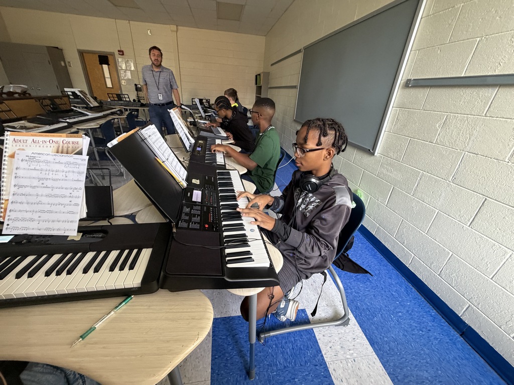 Percussion students are in a classroom with piano keyboards on desks. Their hands are resting on the keys while looking at their band instructor who is standing in the middle of a u-shaped row of desks sharing instructions.