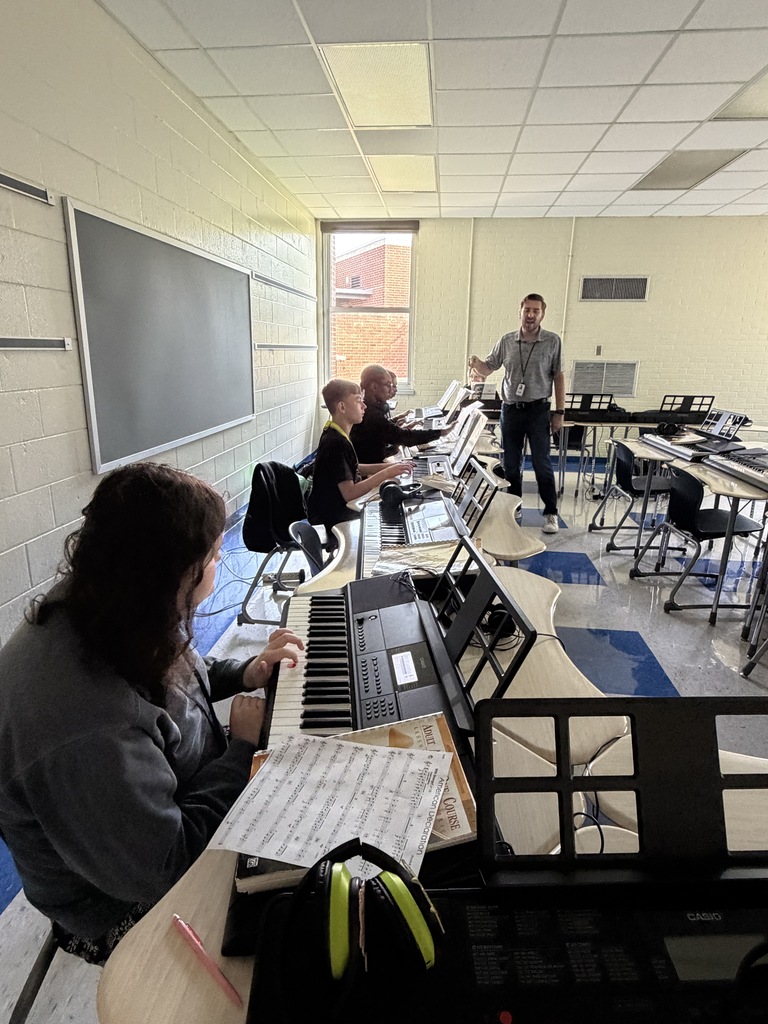 The band instructor is shown walking towards a few students as the class is practicing on the piano keyboard.