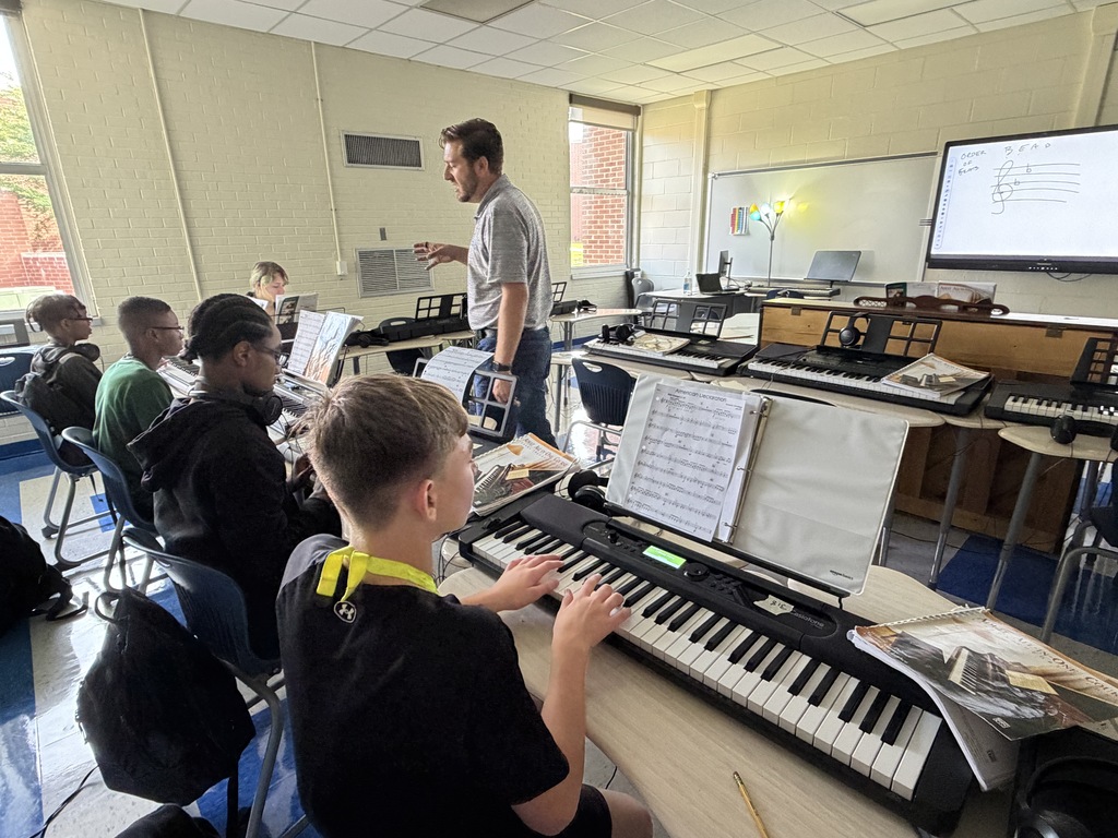 The band instructor is shown walking towards a few students as the class is practicing on the piano keyboard.