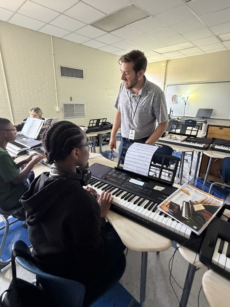 The band instructor is standing in front of a student's sheet music while the student is seated at his desk with the piano keyboard on his desk. The teacher is sharing information with the class about certain notes on the the sheet music.