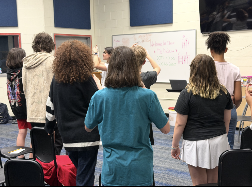 A group of students are standing on risers facing away from the camera and looking at their teacher holding their hands in the air practicing Solfège hand signs, rhythm counting, and vocal warm-ups! 