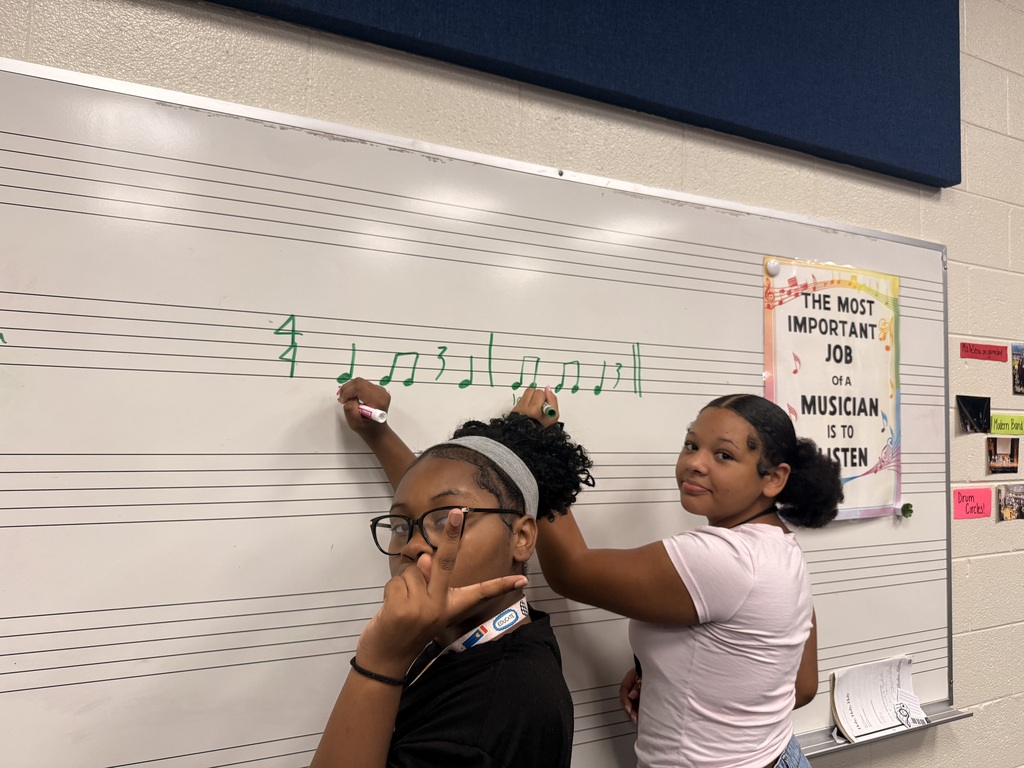 Two femal students are writing on the board different music notes. They both have their hands leaning against the board on with a dry erase marker in their hands. They both are looking at the camera. One is holding a peace sign in the air.