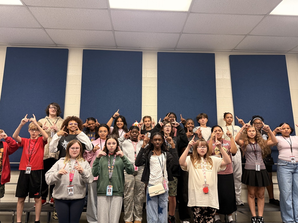 A group of students are standing on risers facing the camera and holding their hands in the air practicing Solfège hand signs, rhythm counting, and vocal warm-ups!