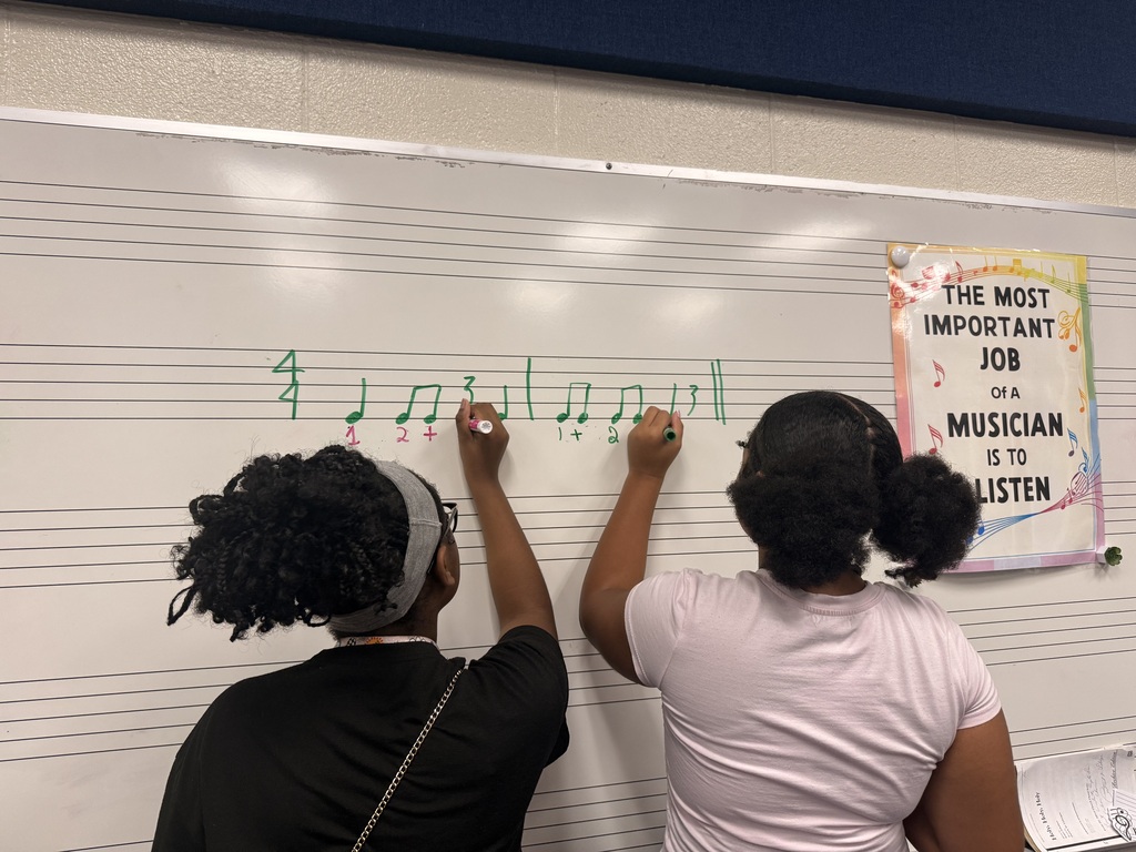 wo female students are writing on the board different music notes. Their backs are turned towards the camera as they write on the board. There is a poster hung on the board to the right of the two students and it states, "The Most Important Job of a Musician is to Listen."