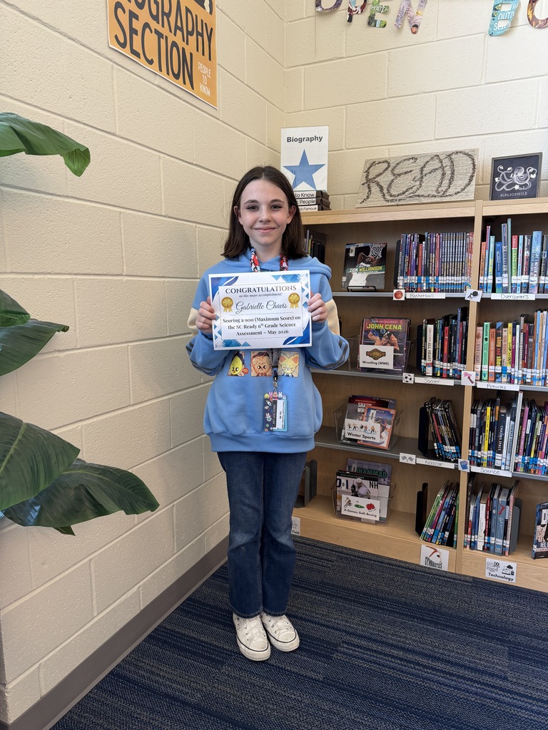 7th grader Gabrielle Chavis is standing in the library near the biography section holding a certificate indicating her accomplishment in receiving a maximum score of a 900 on the SC Ready Science assessment from the 2025-2026 school year.