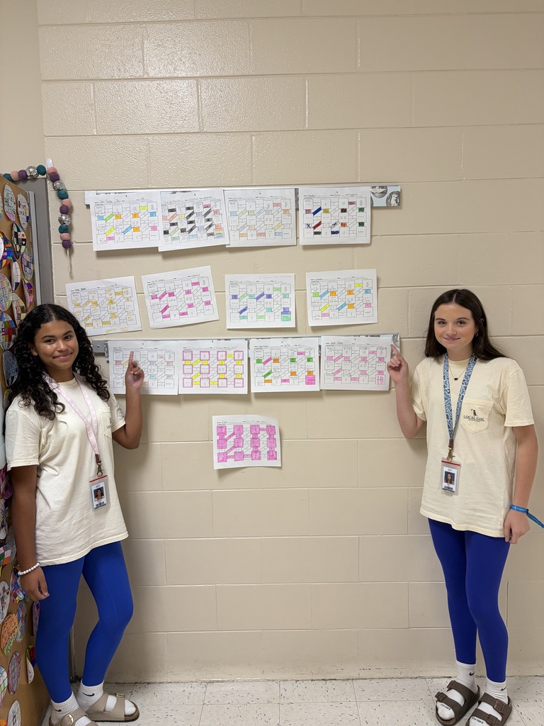 Two female students shown matching in clothing, blue leggings, cream colored t-shirt, white socks, and Birkenstock sandals are shown pointing towards work samples hung on the hallway bulletin board strips outside of their math classroom.