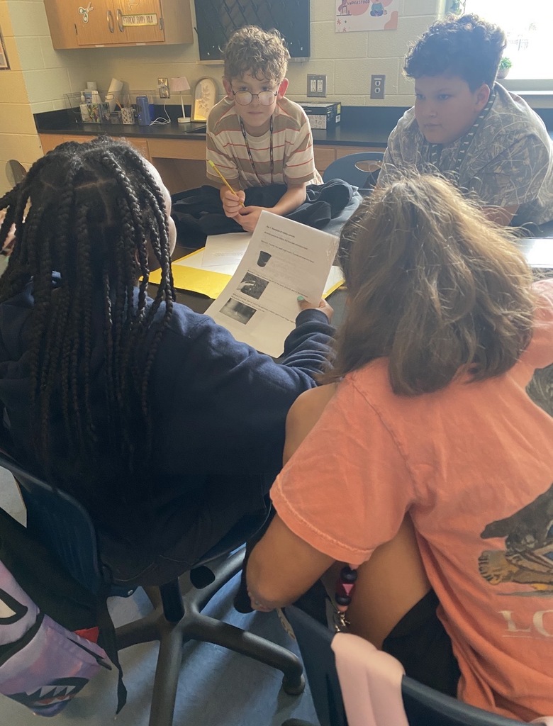 6th grade science students are shown collaborating on an investigation lab while seated and standing at their classroom science lab tables. They are shown looking at and reading paperwork on the tables.