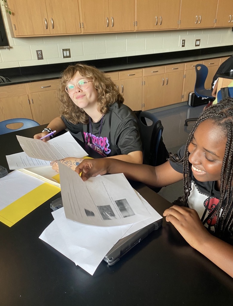 6th grade science students are shown collaborating on an investigation lab while seated and standing at their classroom science lab tables. They are shown looking at and reading paperwork on the tables.