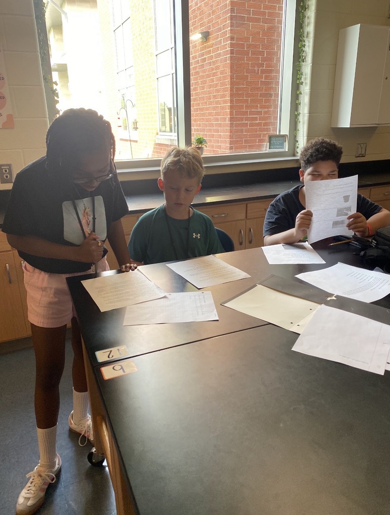 6th grade science students are shown collaborating on an investigation lab while seated and standing at their classroom science lab tables. They are shown looking at and reading paperwork on the tables.