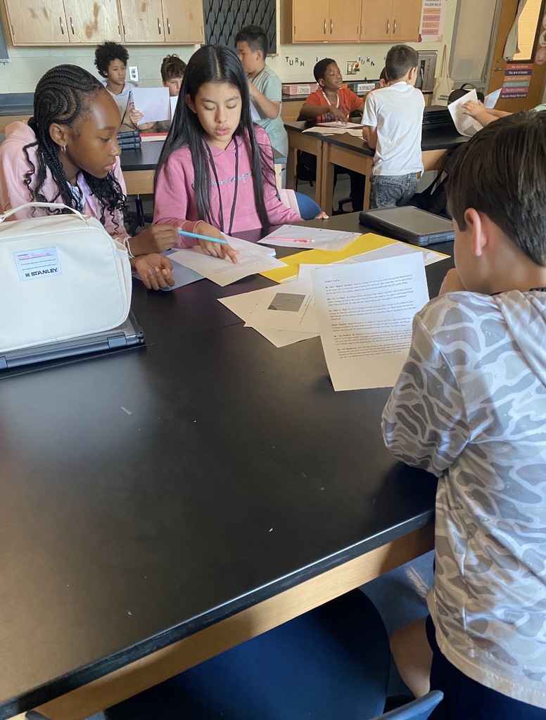 6th grade science students are shown collaborating on an investigation lab while seated and standing at their classroom science lab tables. They are shown looking at and reading paperwork on the tables.