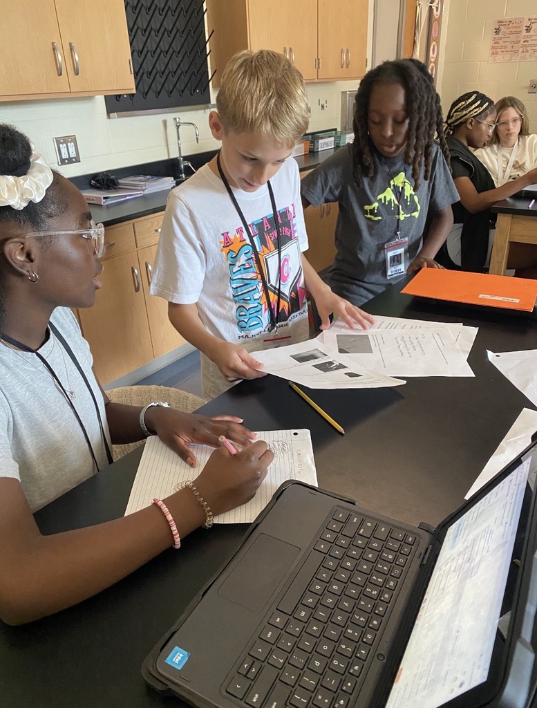 6th grade science students are shown collaborating on an investigation lab while seated and standing at their classroom science lab tables. They are shown looking at and reading paperwork on the tables.