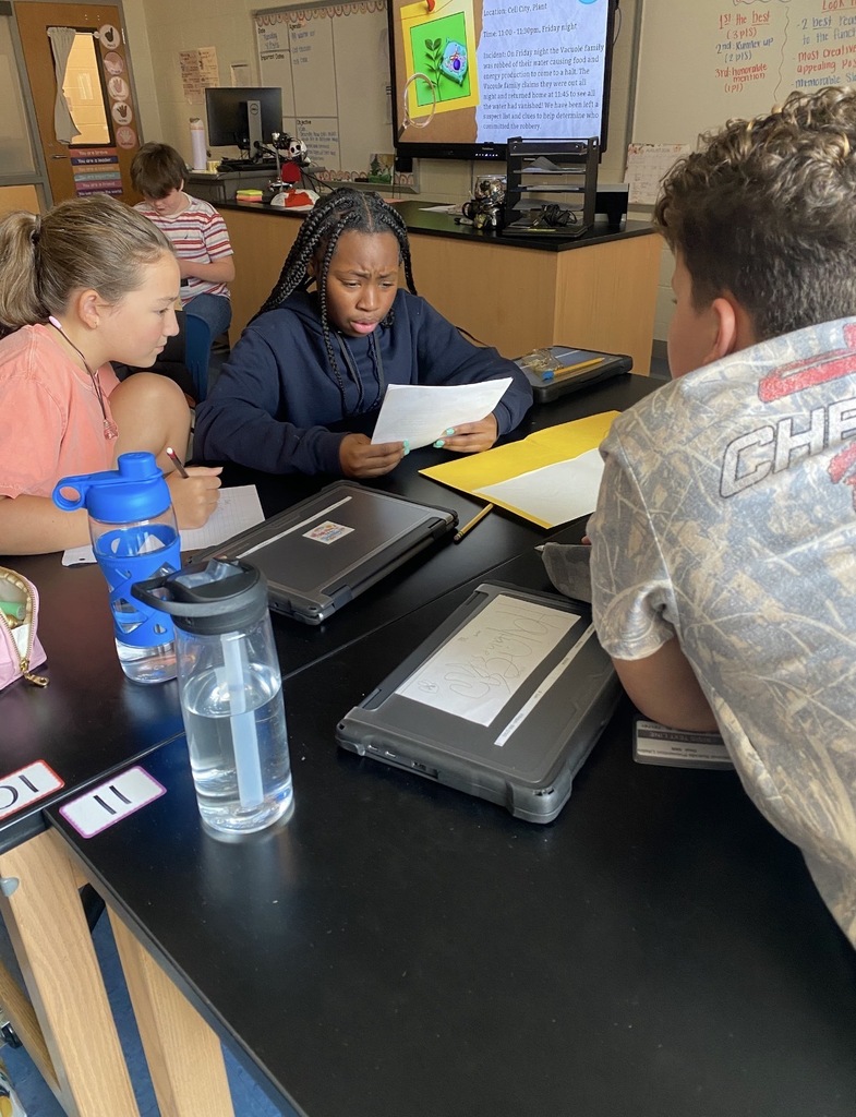 6th grade science students are shown collaborating on an investigation lab while seated and standing at their classroom science lab tables. They are shown looking at and reading paperwork on the tables.