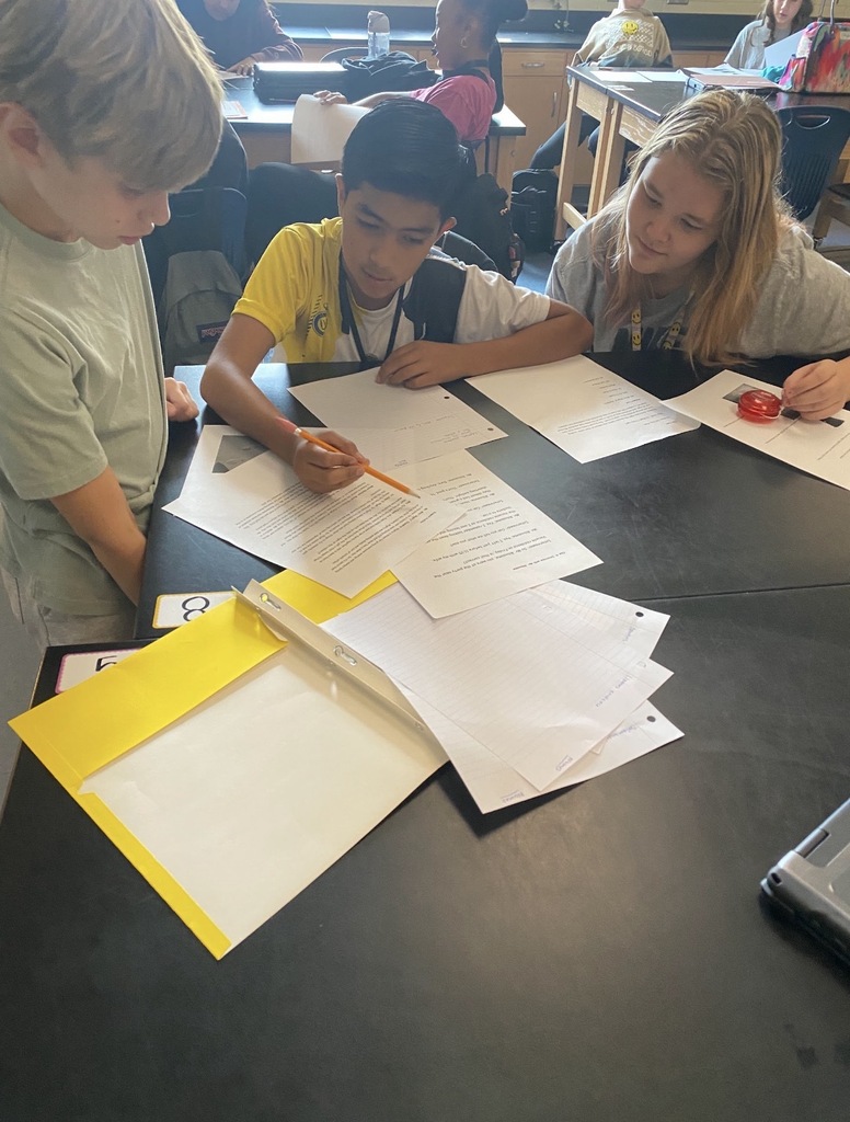 6th grade science students are shown collaborating on an investigation lab while seated and standing at their classroom science lab tables. They are shown looking at and reading paperwork on the tables.