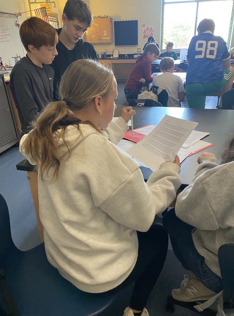 6th grade science students are shown collaborating on an investigation lab while seated and standing at their classroom science lab tables. They are shown looking at and reading paperwork on the tables.