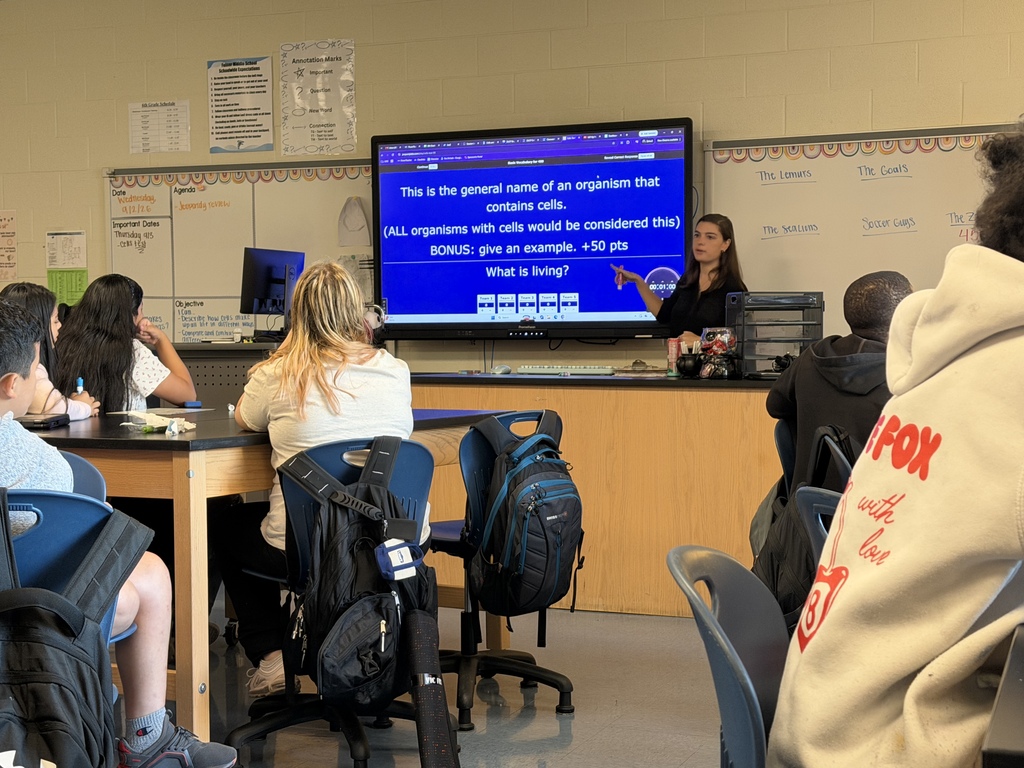 Students are seated at their science lab tables and they are looking at Ms. Lorton who is standing and pointing at her Promethean board as she is going over a question related to cells and living organisms.