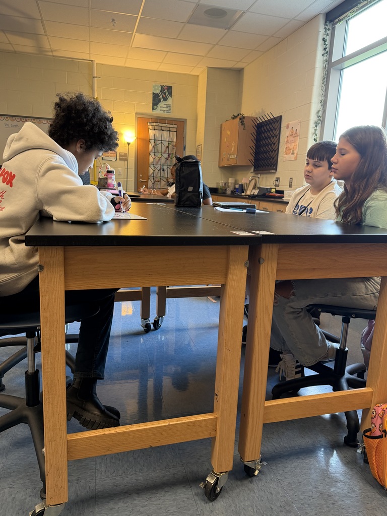 Students are seated at their science lab tables. Some are shown listening to the questions being asked and others are writing something down on a piece of paper.