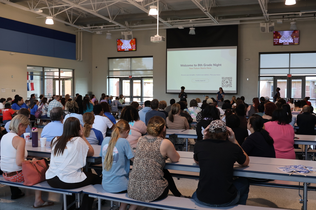A large group of parents/guardians and 8th grade students are shown sitting at cafeteria tables during the 8th Grade Information Night hosted by the School Counseling Department. Two employees are shown at the front of the cafeteria standing in front of the projector screen that shows the heading, "Welcome to 8th Grade Night" and a QR code is shown on the bottom right corner. There is other information below the title, but it is not readable.