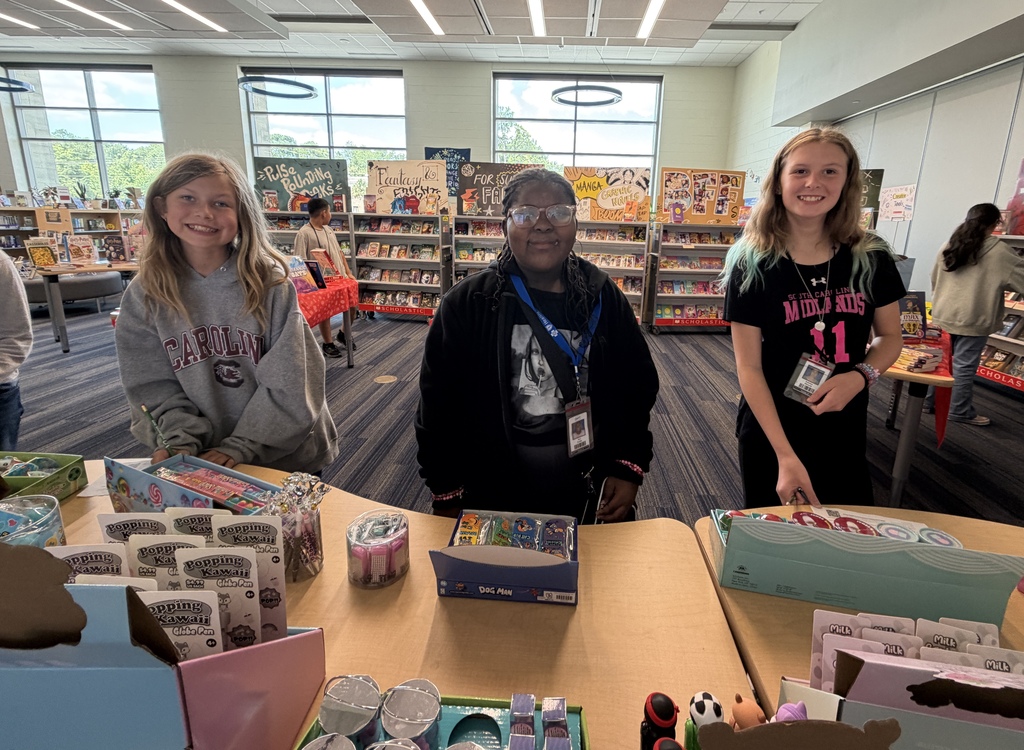 Three female students are standing at the tables in the library while shopping non-book related items at the book fair; they are all smiling at the camera.