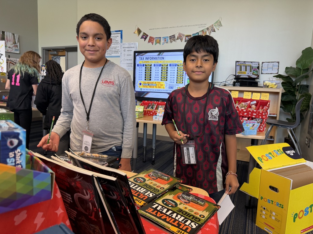 Two male students are smiling at the camera and holding a pencil and paper in their hand as they browse the bookfair.