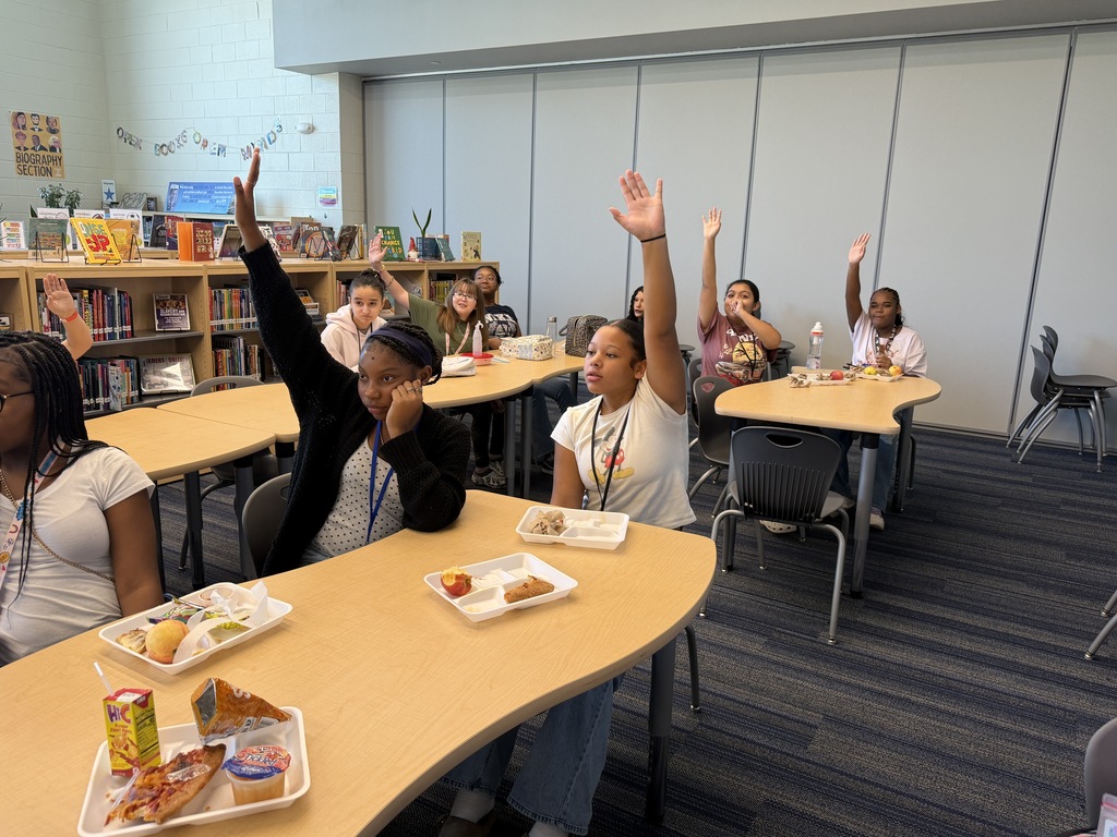 Students gathered in the library sitting at tables while eating lunch and participating in the Fulmer library and public library collaboration event. Students are raising their hands to share a part of speech in a library Mad Lib activity.
