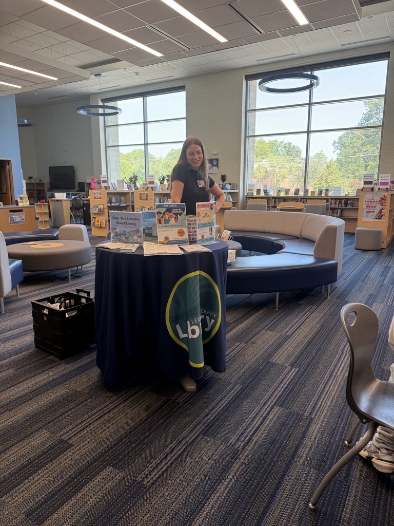 The youth services librarian is standing near a table in the library. The table has the public library table cloth on it with the library emblem. There are library signage stands on the table and other papers with programming information on them and library applications for students to pick up and take home to show their parents or guardians.