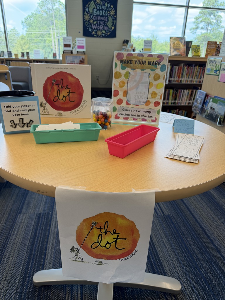 A tall table is shown in the picture containing the book The Dot by Peter H. Reynolds, a sign that instructs students to vote on how many "dots" they think are in the jar shown on the table. The jar is filled with small multi-colored gumballs. There are baskets of voting paper, pencils, a free Dot Day bookmark, and a cup for students to place their guesses in. There is a sign taped to the front of the table that shows a picture of the front of the book.