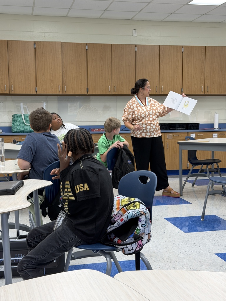 Mrs. Caughman is standing in a classroom while students are seated at their desks listening to her read the picture book The Dot by Peter H. Reynolds in celebration of International Dot Day.