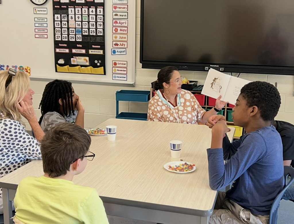 Mrs. Caughman is sitting at a table with another teacher and students in a classroom while the students and teacher are listening to Mrs. Caughman read the picture book The Dot by Peter H. Reynolds in celebration of International Dot Day.