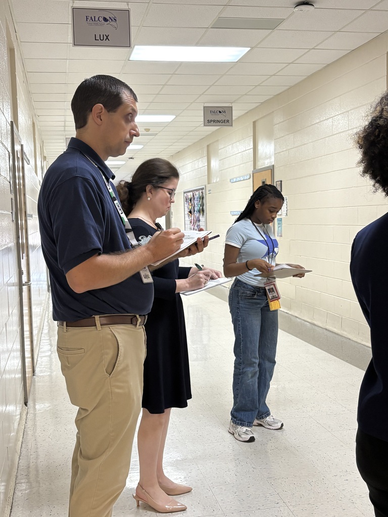 Two adults and a student are shown standing in the hallway holding a paper and pen in their hands assessing college decorated doors done by employees.
