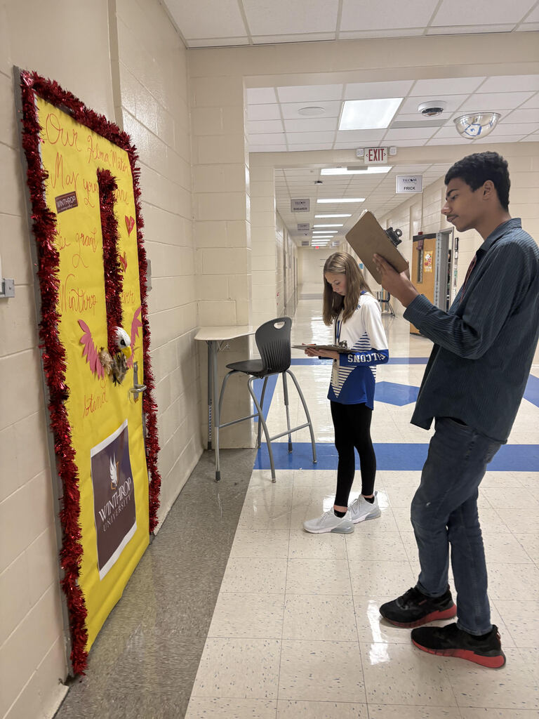 Two students are shown standing in the hallway holding a paper and pen in their hands assessing college decorated doors done by employees.