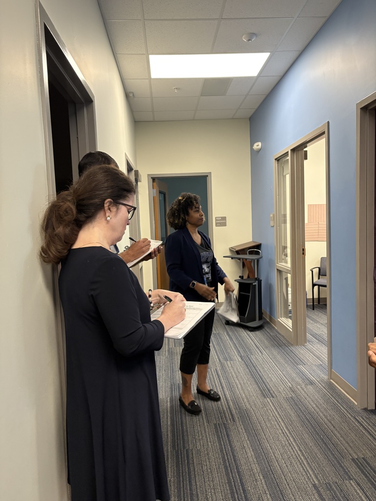 Two adults are shown standing in the hallway holding a paper and pen in their hands assessing college decorated doors done by employees. The school social worker is also shown standing near the adults.