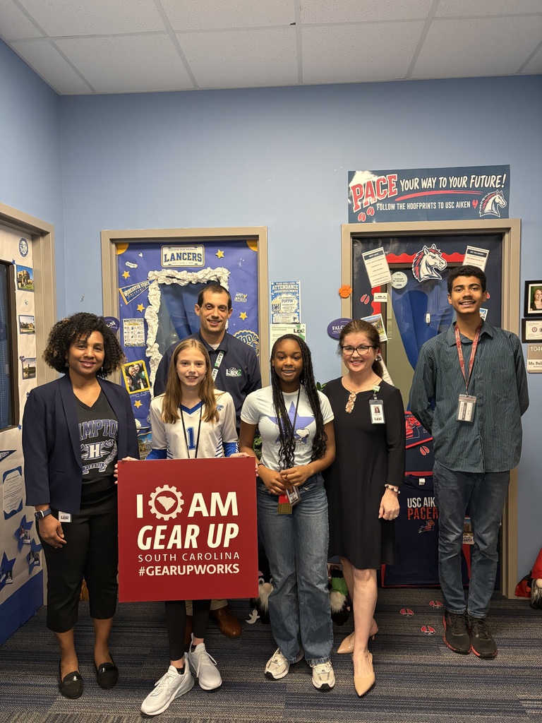 The adults, the school social worker, and the students who assessed and voted on the different college decorated doors are standing and smiling for the camera. One of the students in the front is hold a square red sign that states, "I am Gear Up, South Carolina #GearUpWorks.