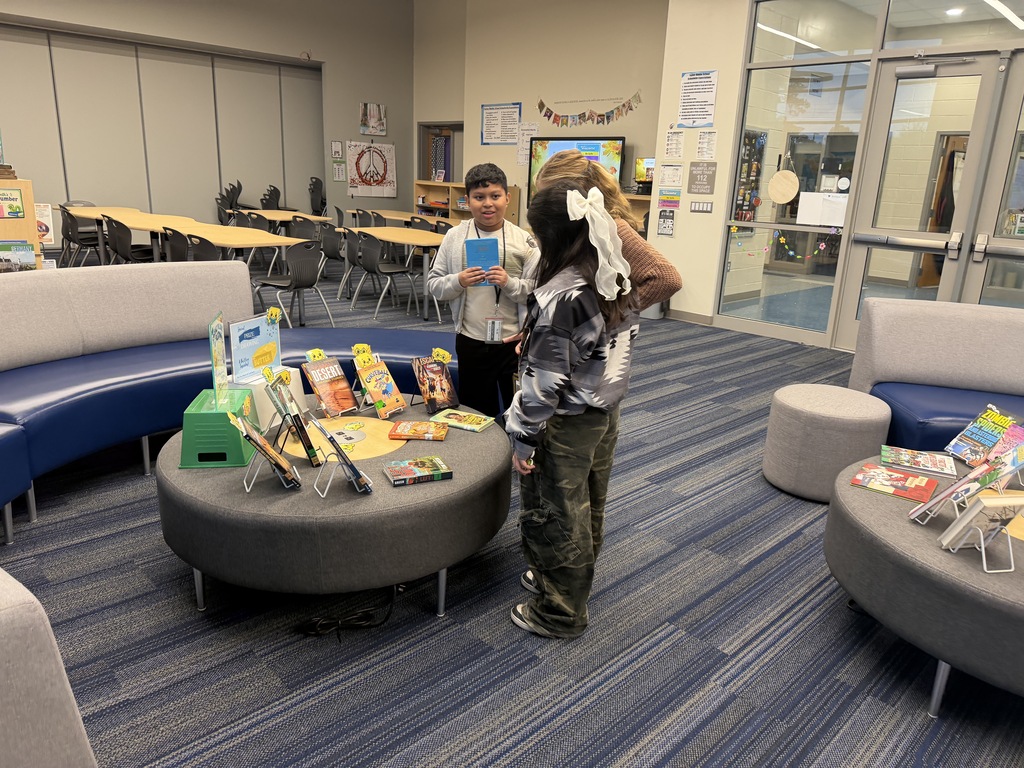 Three students are shown standing next to the "Butter Book Display" table in the library next to the couches. This display has books on and off of easels. There are slices over butter on "book peepers" peeking out of some of the books. The display sign about "Spread the Love of Reading" and "You Better Believe these are google books" are in clear acrylic stands sitting on top of upside down storage bins so students can view the signage. The students are discussing the books and the new reading challenge.