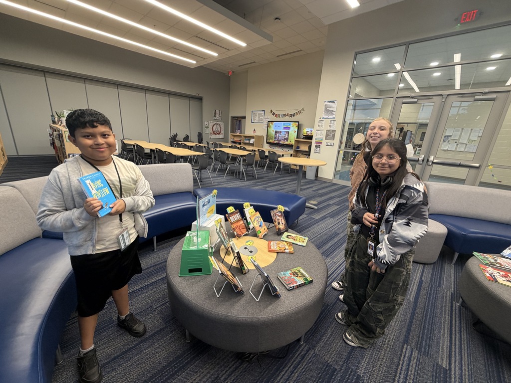 Three students are shown standing next to the "Butter Book Display" table in the library next to the couches. This display has books on and off of easels. There are slices over butter on "book peepers" peeking out of some of the books. The display sign about "Spread the Love of Reading" and "You Better Believe these are google books" are in clear acrylic stands sitting on top of upside down storage bins so students can view the signage. The students are looking at the camera and smiling.
