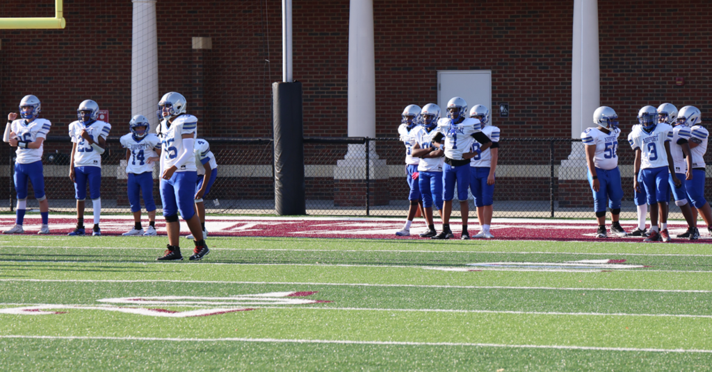 The C-team football team is shown warming up prior to the game. They are standing in separate small groups in the endzone with one player on the 5 yard line.