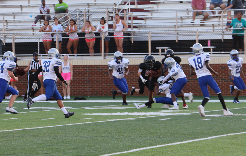 The football players are shown running on the field with one of the Airport C-team players almost on the ground as he grabs the opponents legs.