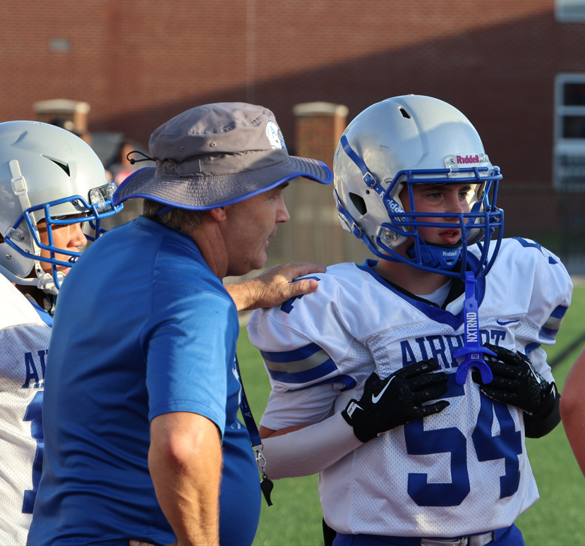 A C-team football coach is shown with his hand on the shoulder pad of a player who is looking off into the field. The coach is talking with the player as the player listens. There is another football player shown on the left side of the coach standing and watching the field.