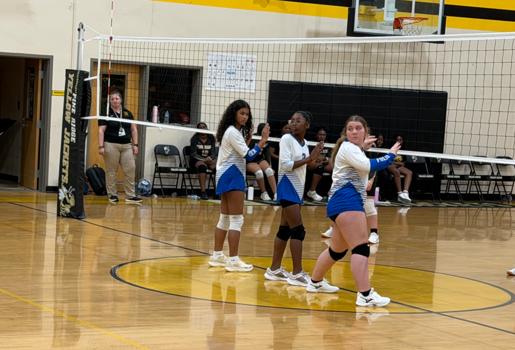 Three female volleyball players are standing at the volleyball net looking backwards waiting on their teammate to serve.