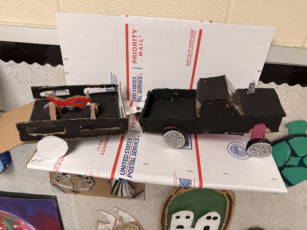 A black truck is laying on a folded piece of cardboard. It is pulling a cardboard trailer that is carrying a dirt bike. All made out of cardboard.
