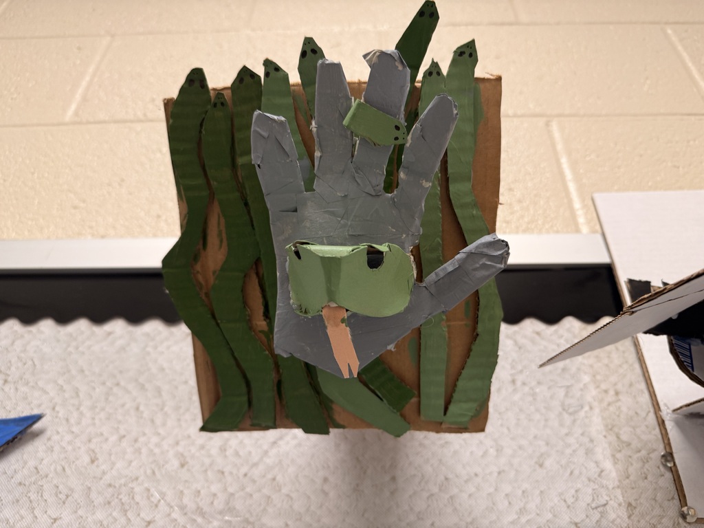 A 3D cardboard design with green colored snakes vertically displayed upward on a brown cardboard piece. Tere is a hand cardboard coming out from the background in the color of gray with a snake head wrapped around the middle finger and a snake's face coming outward from the palm of the hand.
