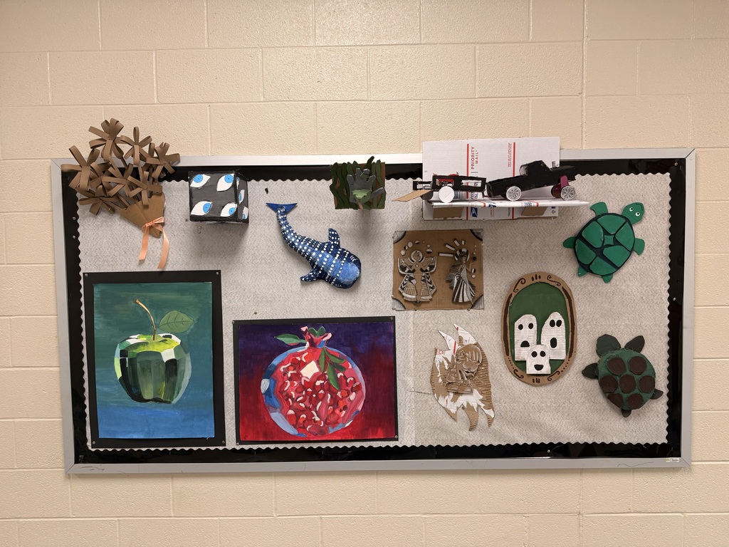A bulletin board display of various 3D artwork designs and some illustrated paintings of fruit. Some of the 3D designs include: two turtles, a frame with three white ghosts, a truck pulling a trailer holding a dirt bike, a hammerhead shark, and a bouquet of flowers.
