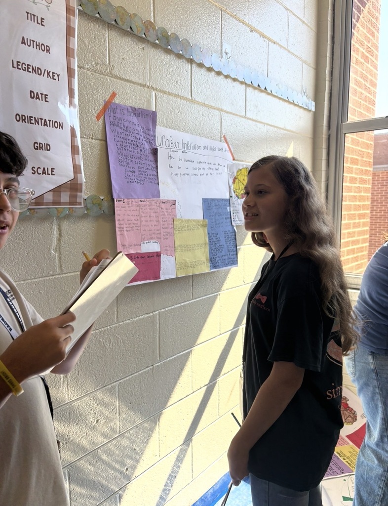 7th grade social studies students are conducting a gallery walk by viewing different student group posters that share information about West African culture as they wrapped up this unit in class. Students are shown walking around with a paper and pencil in hand as they look at the different posters taped on the wall in different places in the classroom.