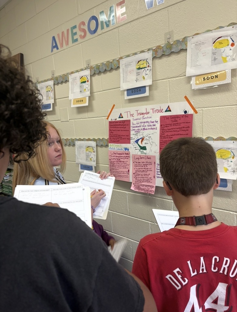 7th grade social studies students are conducting a gallery walk by viewing different student group posters that share information about West African culture as they wrapped up this unit in class. Students are shown walking around with a paper and pencil in hand as they look at the different posters taped on the wall in different places in the classroom.