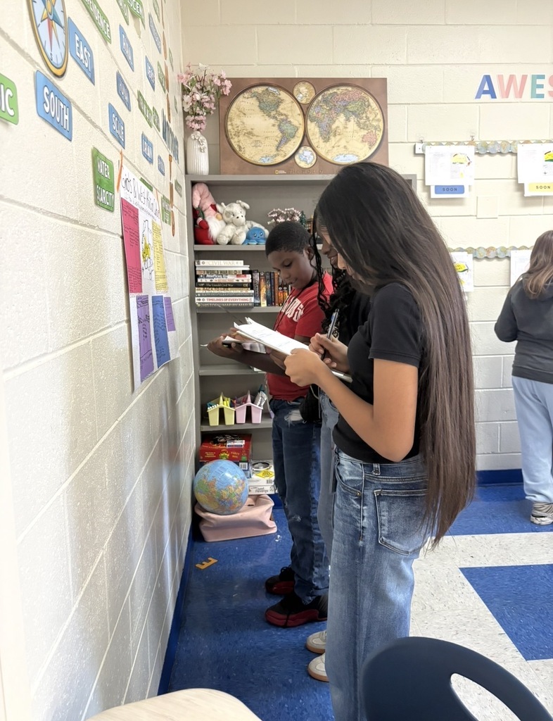 7th grade social studies students are conducting a gallery walk by viewing different student group posters that share information about West African culture as they wrapped up this unit in class. Students are shown walking around with a paper and pencil in hand as they look at the different posters taped on the wall in different places in the classroom.
